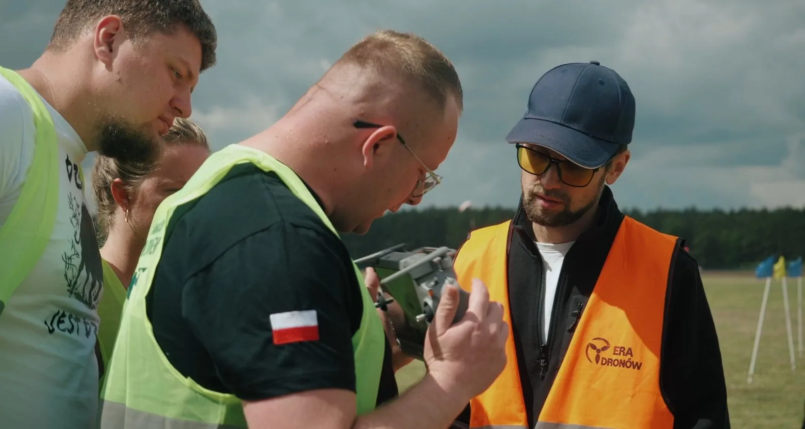 Instruktor Era Dronów w pomarańczowej kamizelce pomaga uczestnikowi szkolenia obsługiwać aparaturę drona