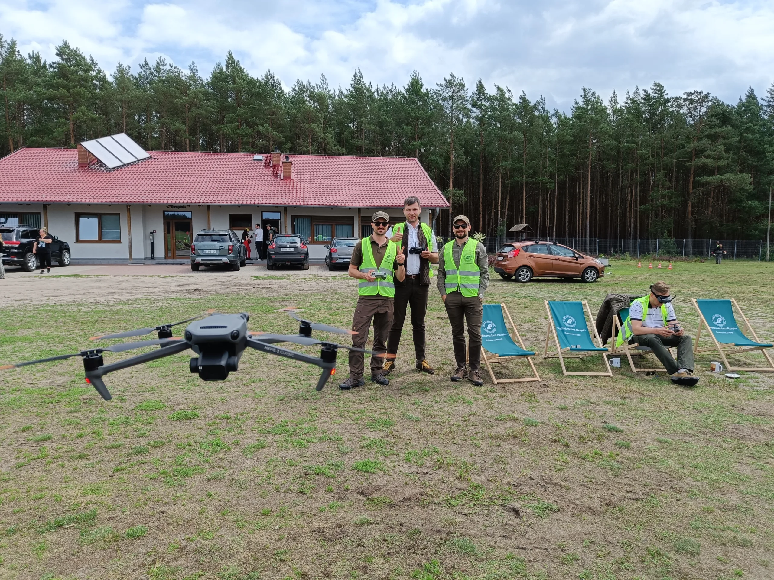 Kursanci w kamizelkach odblaskowych podczas lotów praktycznych na szkoleniu Era Dronów — dron Mavic 3 Classic w zawisie nad lądowiskiem